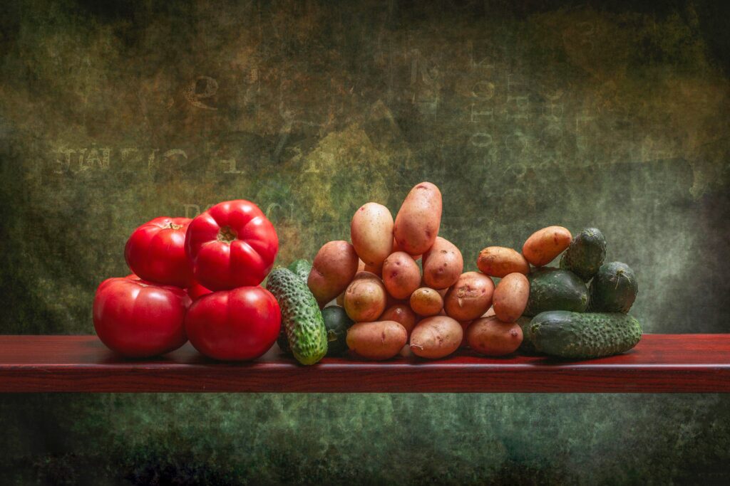 A rustic display of fresh tomatoes, potatoes, and cucumbers on a wooden shelf against a textured background.